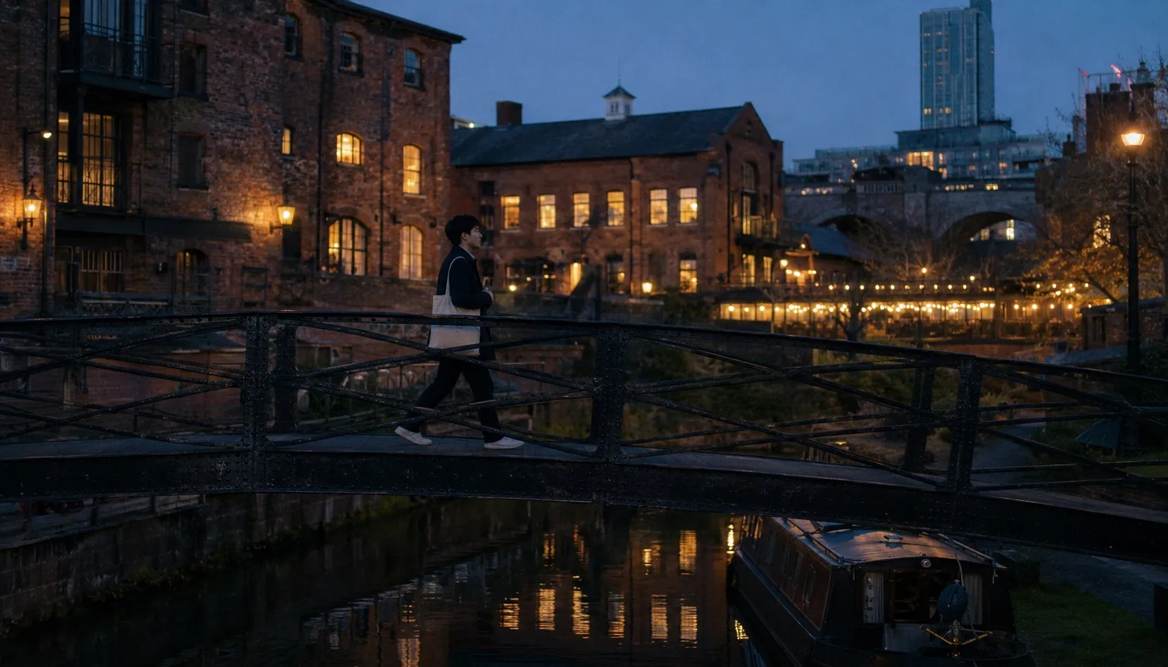 A single student walking across an iron footbridge over the Castlefield canal at blue hour, red-brick Victorian warehouses lit warm in the background, the Beetham Tower in the distance, a narrowboat moored below